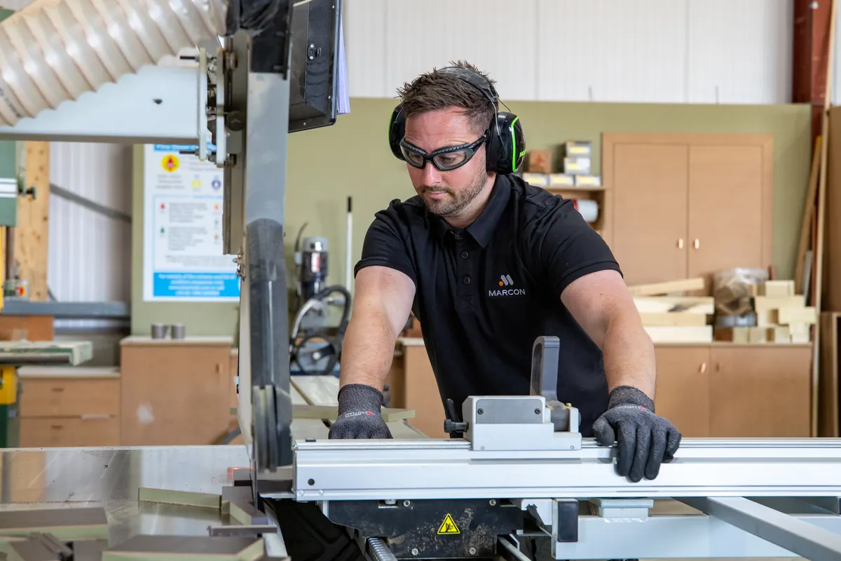 A Marcon joiner in ear defenders, safety glasses and gloves cutting a panel on the panel saw in the joinery workshop